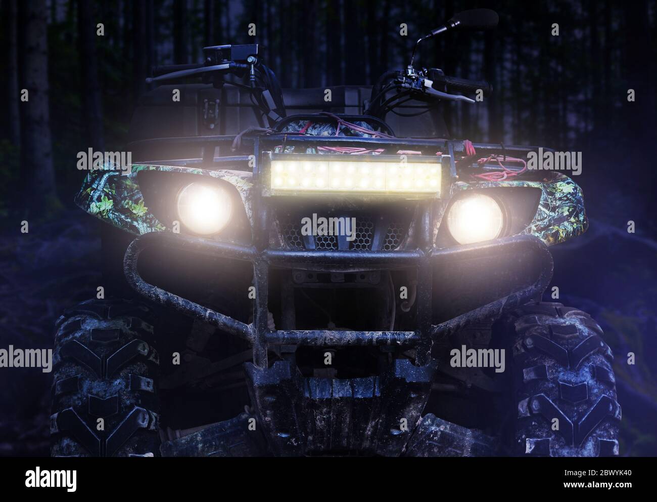 Photo d'un véhicule tout-terrain de chasse sale et camouflé debout dans la forêt la nuit avec les phares allumés, vue avant. Banque D'Images