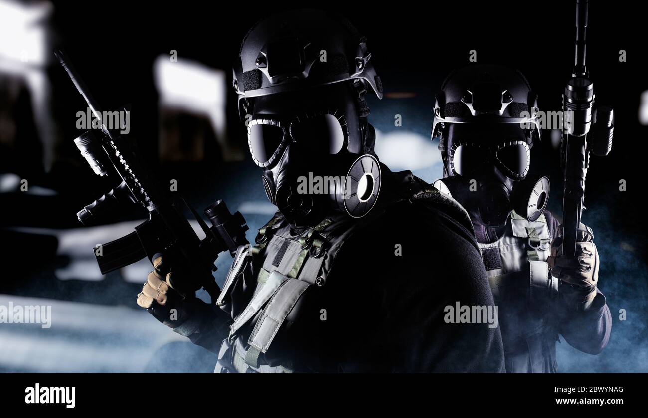 Photo d'un militaire entièrement équipé en gilet tactique d'armure noire, masque à gaz, fusil automatique, gants et casque debout sur fond de hall sombre. Banque D'Images