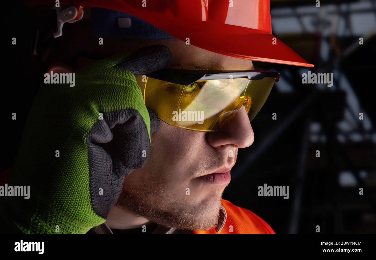 Photo d'un ouvrier en costume de veste orange avec des gants de protection et un casque mettant des lunettes sur le fond de la zone de construction. Banque D'Images