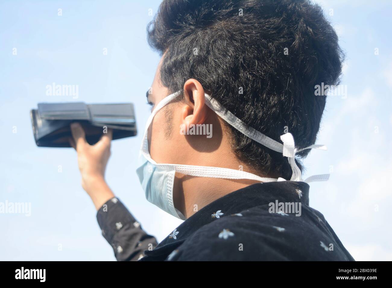 Jeune homme portant un masque facial de protection et montrant son portefeuille vide sur fond de nuages et de ciel. Banque D'Images