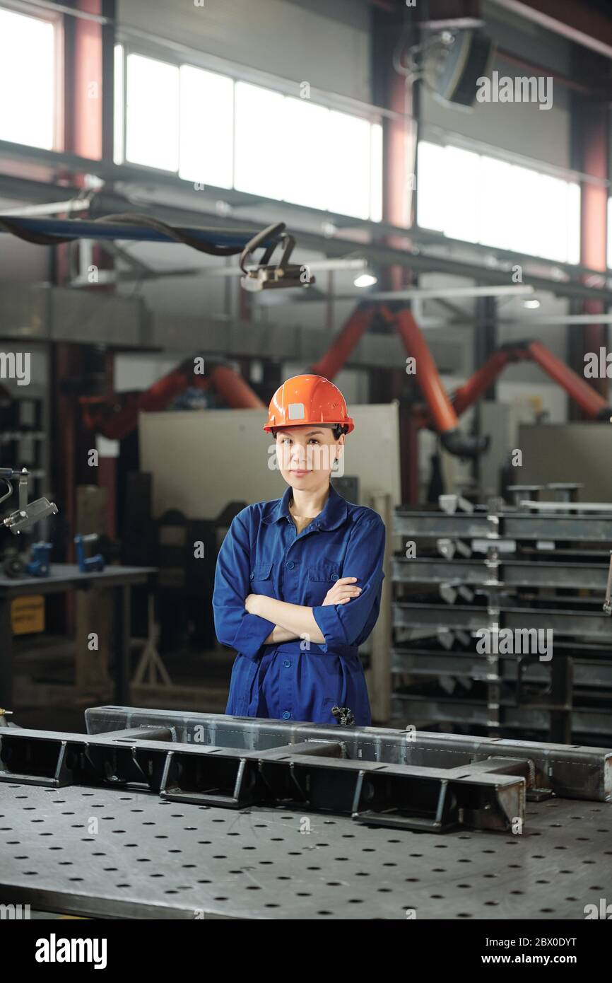 Portrait du contenu attrayant spécialiste asiatique du travail des métaux en casque debout à table avec des détails métalliques dans l'atelier Banque D'Images