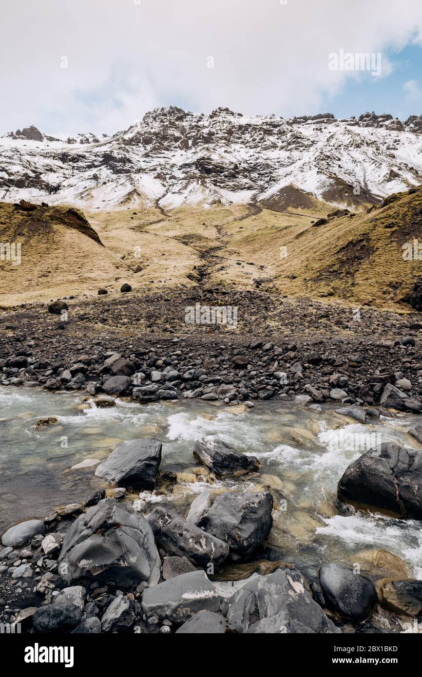 Rivière de montagne au pied de la montagne avec un sommet enneigé. Herbe jaune sèche sur les montagnes en mai en Islande. Banque D'Images