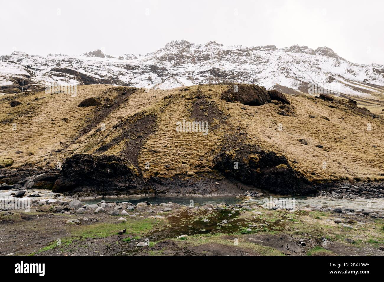 Rivière de montagne au pied de la montagne avec un sommet enneigé. Herbe jaune sèche sur les montagnes en mai en Islande. Banque D'Images