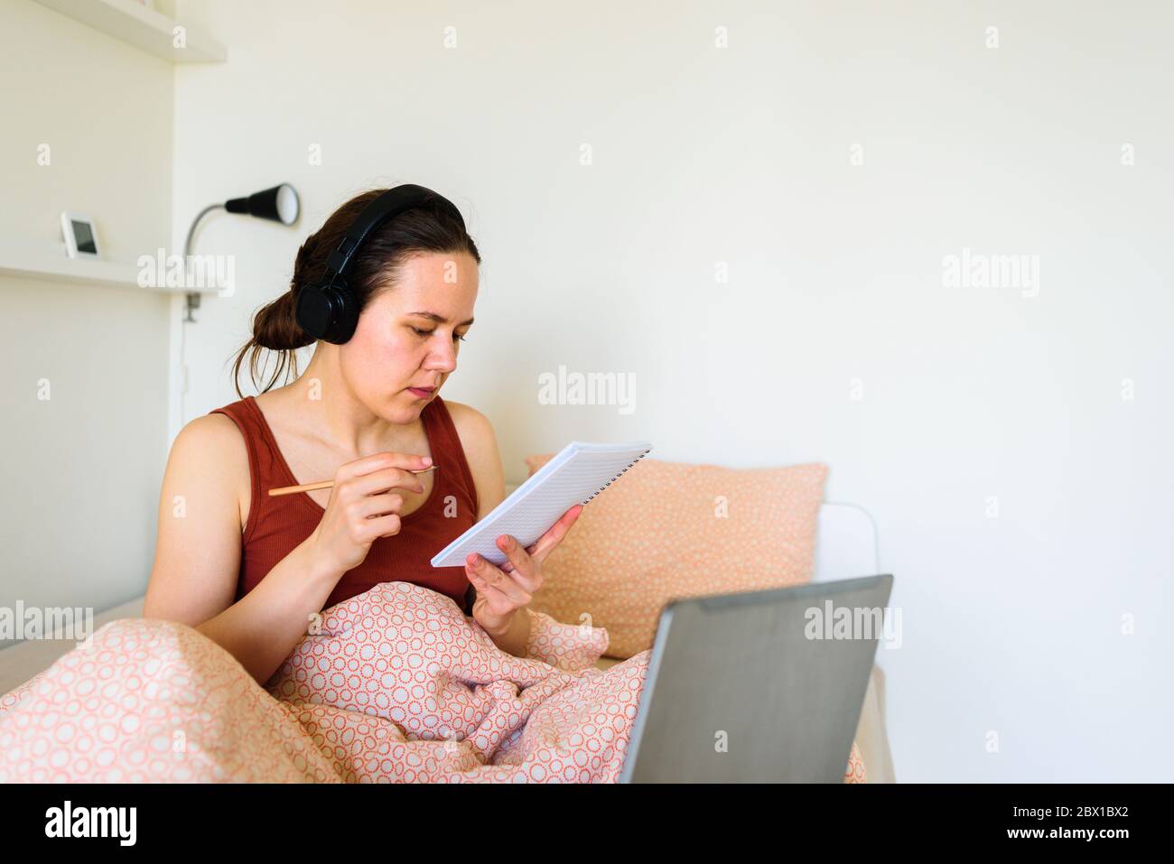 Femme souriante travaillant de la maison dans le lit. Écriture dans le bloc-notes. La vraie vie, le bureau dans la chambre Banque D'Images