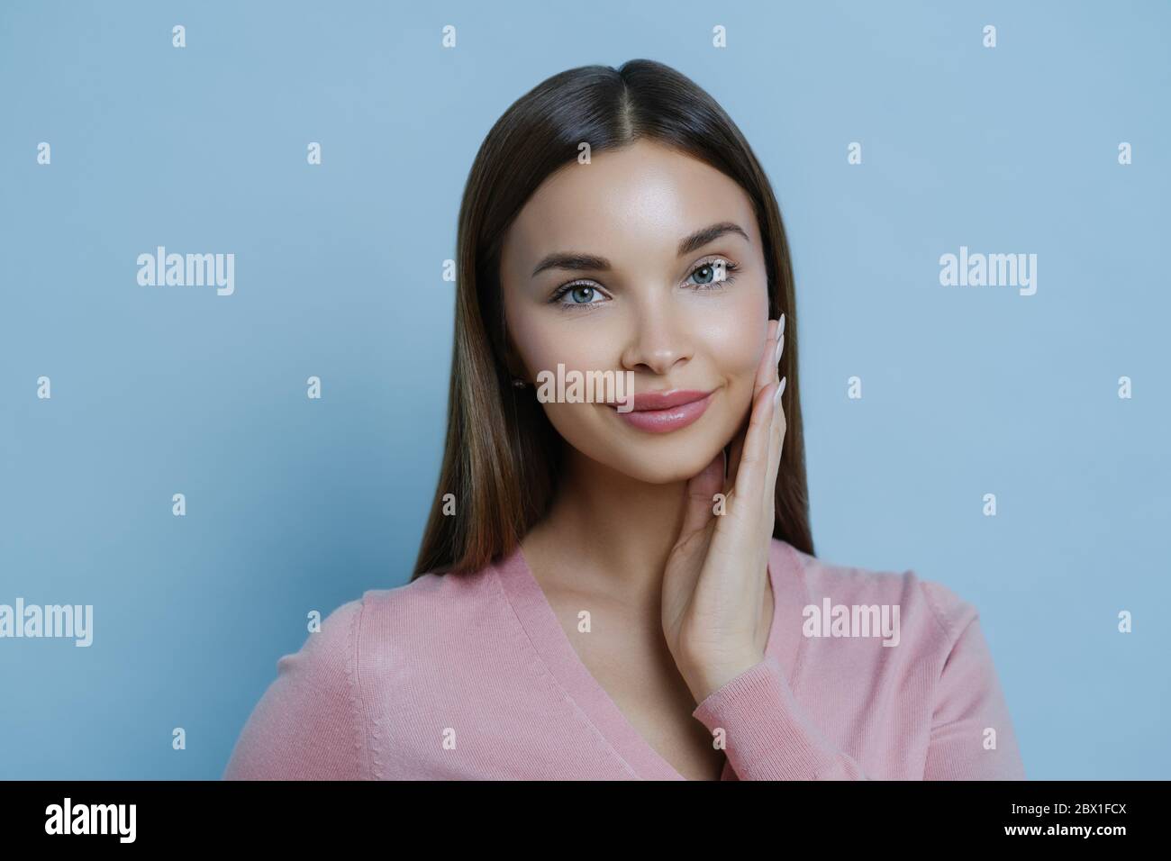 Photo de jeune femme adorable millénaires touche joue doucement, apprécie la douceur de la peau après l'application du masque de beauté ou de la crème pour le visage, regarde à l'appareil photo, iso Banque D'Images