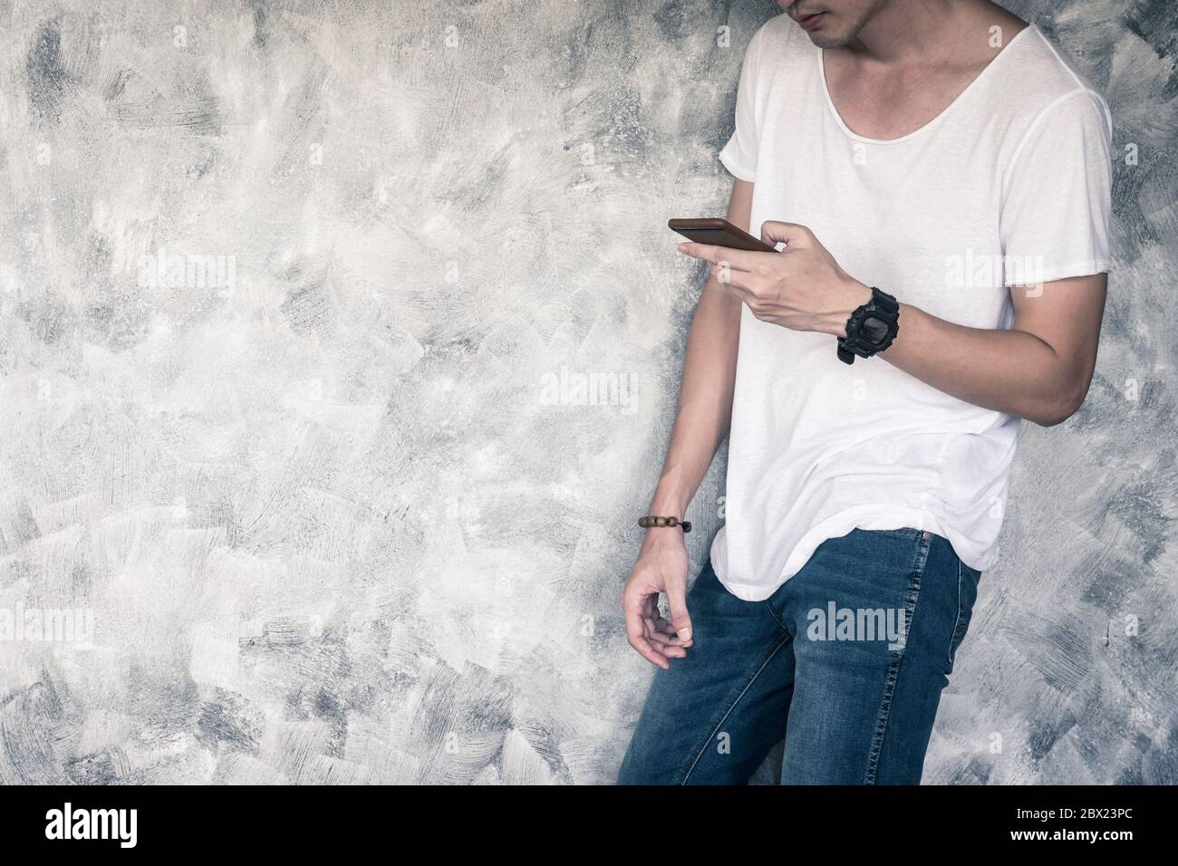 Jeune homme portant un t-shirt blanc et un Jean bleu, tenant un smartphone et debout sur fond gris grunge. Espace libre pour le texte Banque D'Images