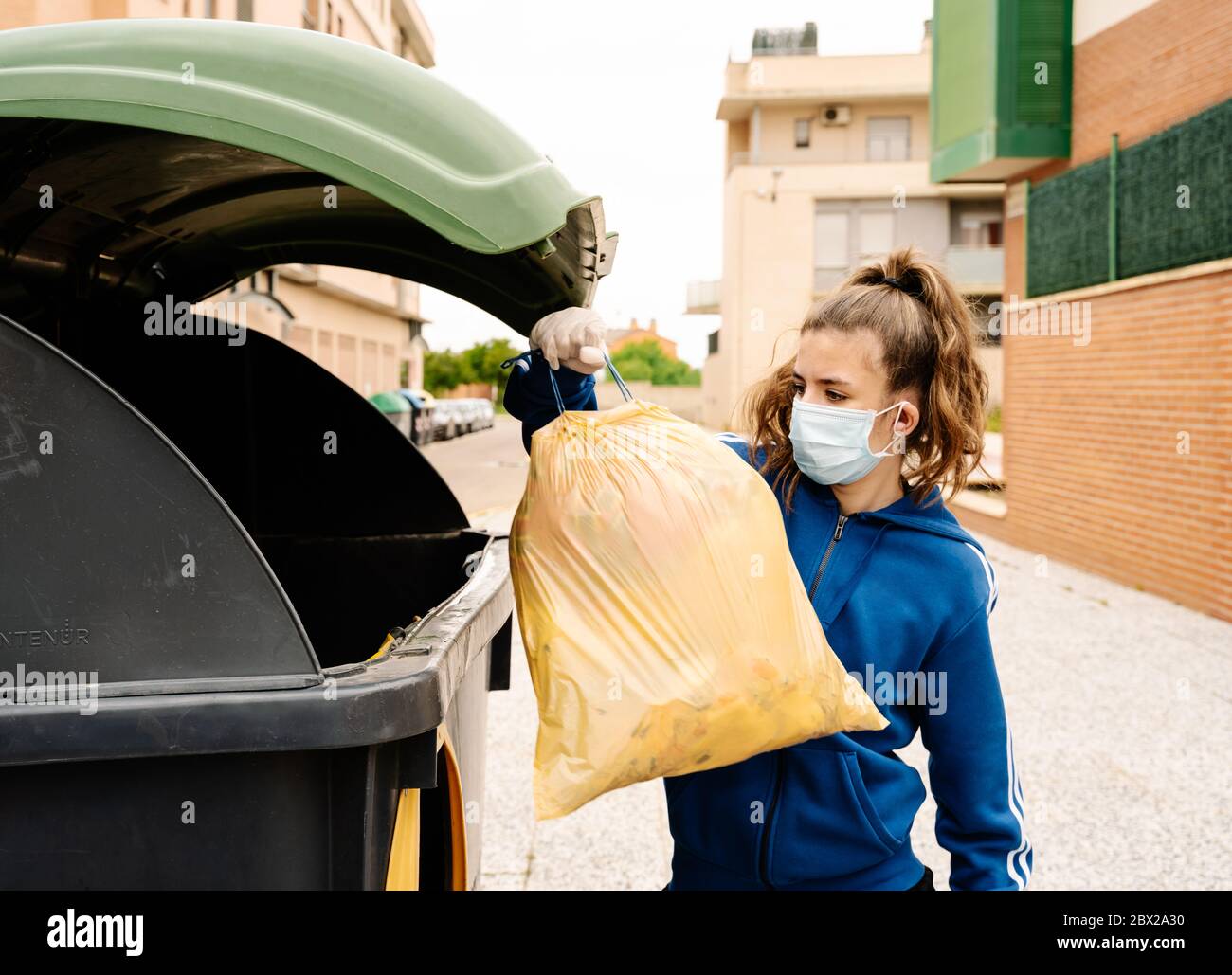 fille jetant un sac poubelle jaune dans un conteneur de recyclage vert  ouvert dans la rue. L'adolescent porte un masque et des gants pour protéger  ses personnes Photo Stock - Alamy