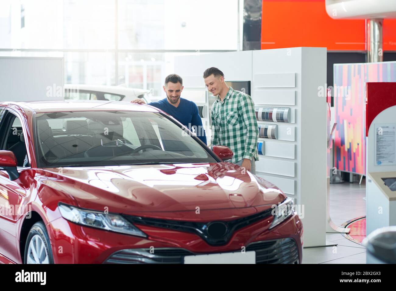 Vendeur de voiture professionnel aidant jeune homme client dans le choix de voiture pour l'achat. Joyeux sourire beau homme dans la chemise regardant la belle voiture rouge avant d'acheter un véhicule réussi. Banque D'Images