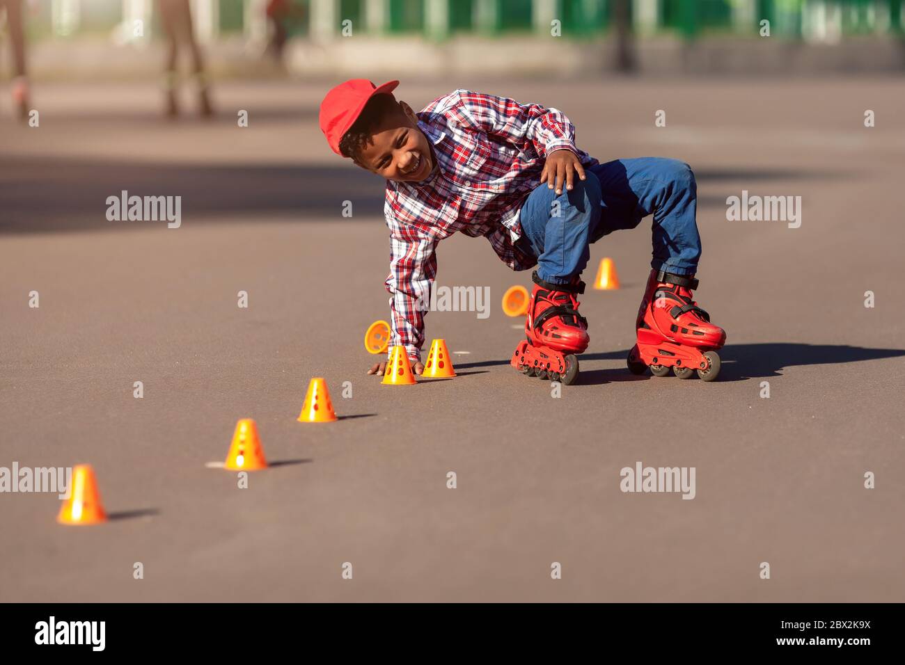 Joyeux petit garçon américain africain sur des patins à roulettes dans le parc d'été sur la route d'asphalte parmi les cônes d'entraînement . Roller enfant est tombé et souriait en essayant de se lever. Banque D'Images