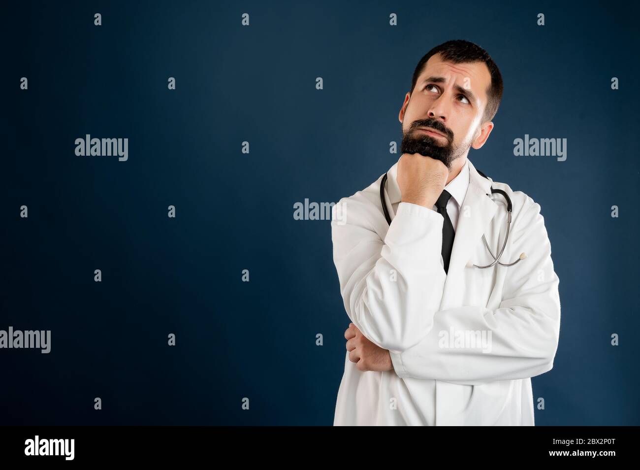 Portrait d'un médecin masculin avec stéthoscope en uniforme médical tenant la main sous son menton, ayant douteux posé sur un fond bleu isolé. Banque D'Images