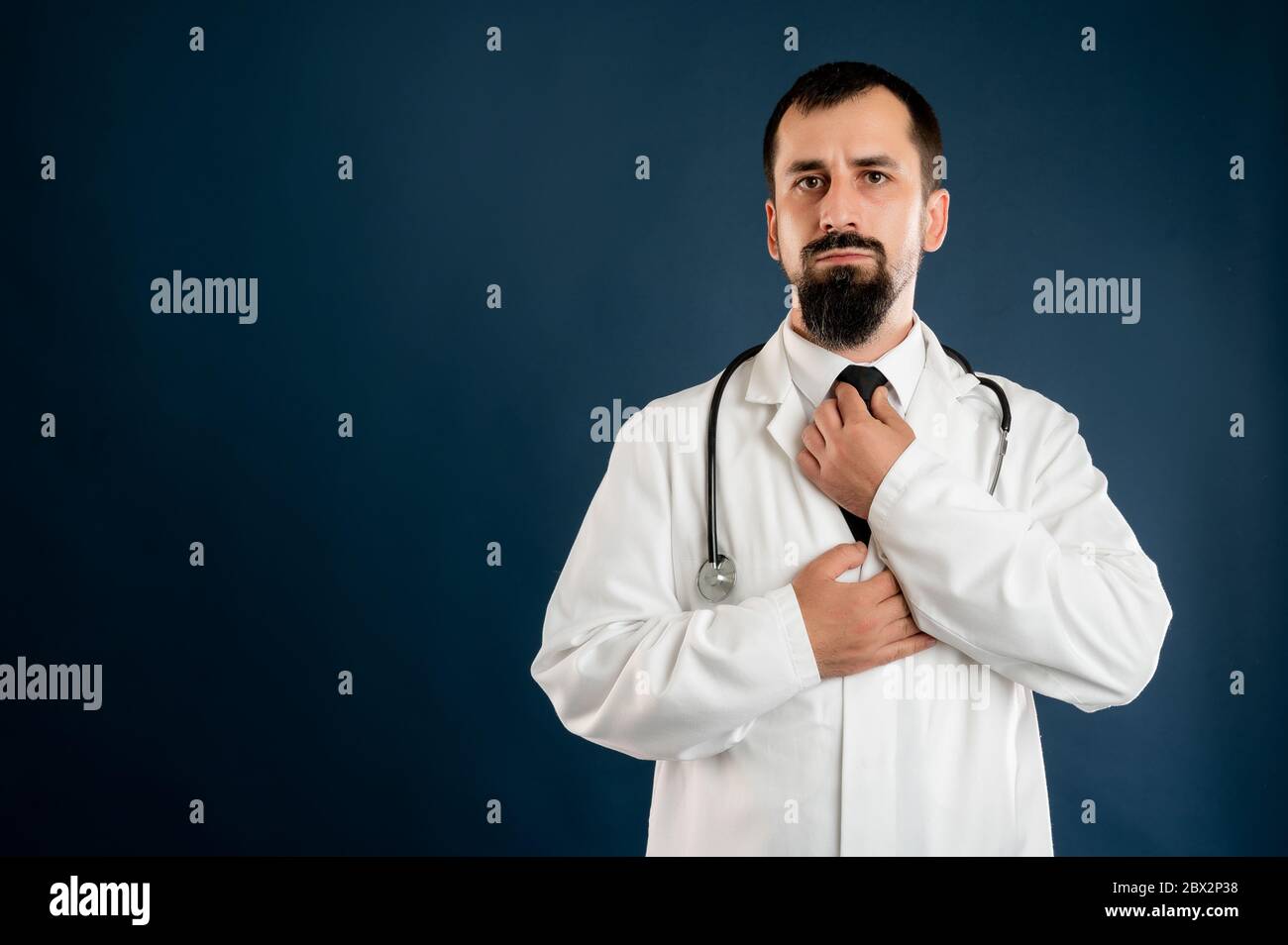 Portrait d'un médecin masculin avec stéthoscope en uniforme médical organise sa cravate posant sur un fond bleu isolé. Banque D'Images