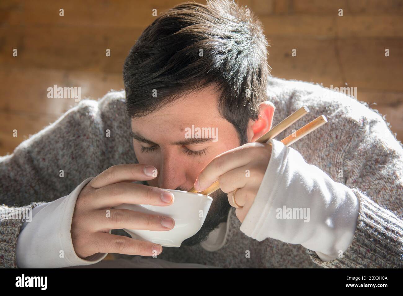 Portrait d'un homme barbu assis et mangeant d'un bol, en utilisant des baguettes, dans une cabine en bois. Banque D'Images