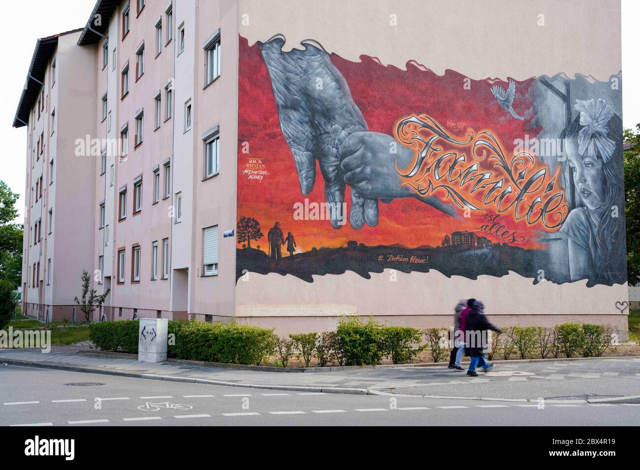 05 juin 2020, Bade-Wurtemberg, Mannheim : trois femmes marchent devant une façade de maison dans le quartier de Schönau avec une fresque de l'artiste graffiti de Mannheim Rick Riojas. Le graffiti montre le motif d'une fille tenant la main d'une personne plus âgée. Le lettrage « Family is Everything » est pulvérisé entre les deux. Les œuvres d'art de Riojas, qui sont appliquées aux maisons de l'association de logement GBG de Mannheim, ne seront visibles que pendant un certain temps jusqu'à ce que les bâtiments soient rénovés. Photo: Uwe Anspach/dpa - ATTENTION: Seulement pour usage éditorial en rapport avec un rapport sur les peintures murales sur les façades de maisons de Mannheimer W Banque D'Images