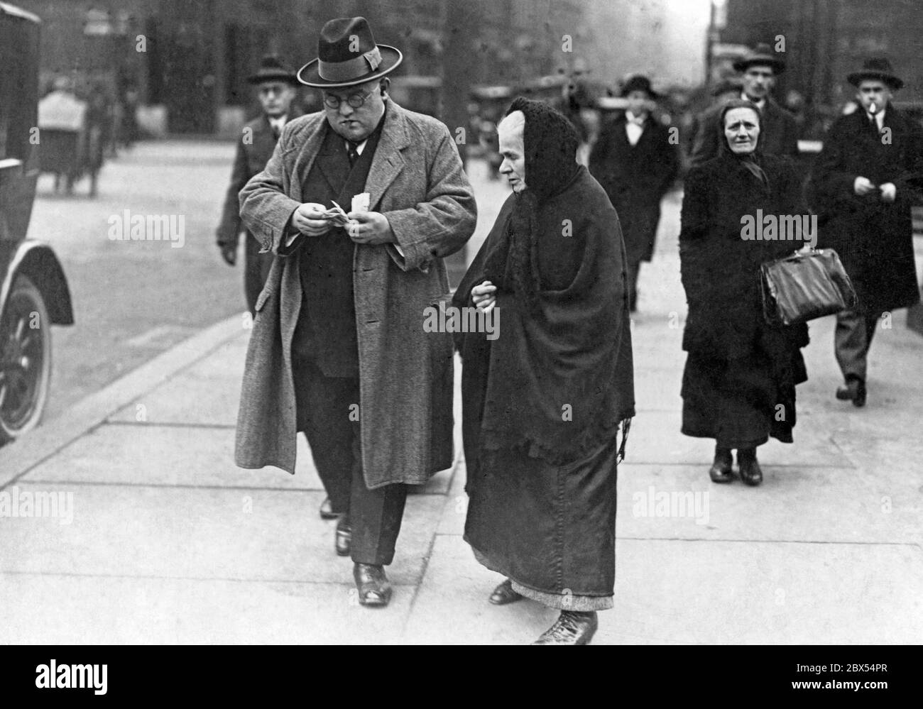Un homme bien habillé portant un manteau et un chapeau donne de l'argent à une vieille femme. La femme porte une jupe noire, une cape noire et un bandana noir. Banque D'Images