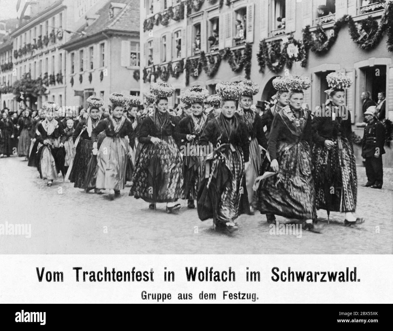 Un groupe de femmes en costume traditionnel marche en procession dans le village décoré de Wolfach dans la Forêt Noire en 1912, portant des costumes de fête et des couronnes de mariée (Schaeppel). Les spectateurs regardent depuis les fenêtres. Banque D'Images