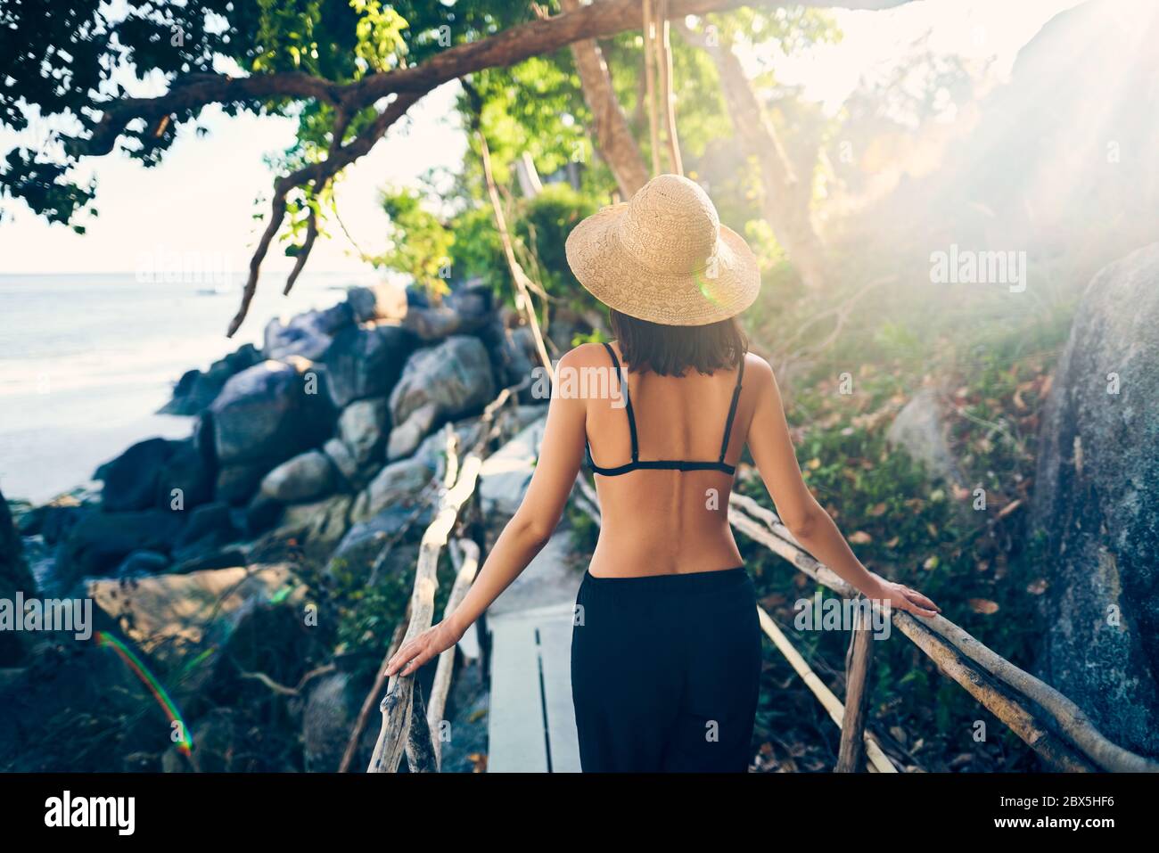 Vue arrière de la jeune femme en chapeau de paille marchant sur un pont en bois dans une île tropicale avec rétroéclairage. Vacances, concept de voyage Banque D'Images