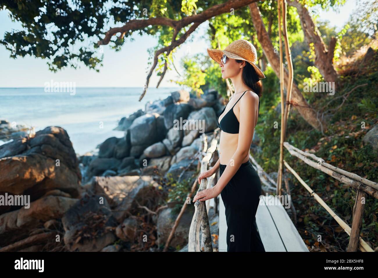 Jeune femme en chapeau de paille profiter de la vue sur la mer sur pont en bois dans l'île tropicale avec rétroéclairage. Vacances, détente, concept de voyage Banque D'Images