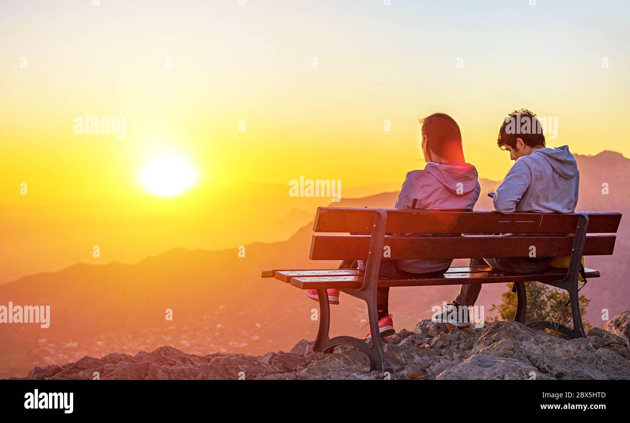 Deux personnes, en couple assis ensemble sur un banc au sommet d'une montagne à la recherche au coucher du soleil au cours de l'heure d'or. Banque D'Images