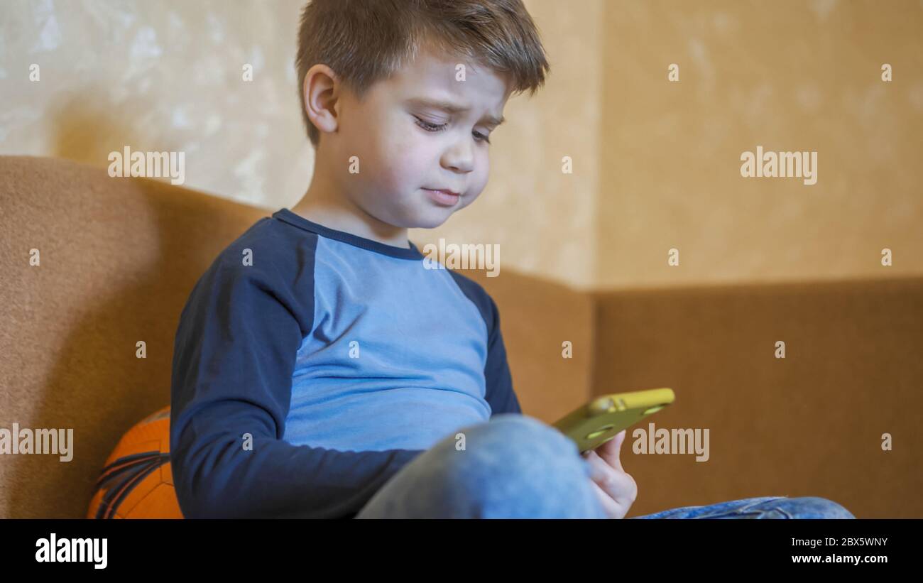 Un garçon de préchooler contrarié par la défaite dans un jeu mobile. Enfant triste utilisant un téléphone portable intelligent assis sur un canapé Banque D'Images