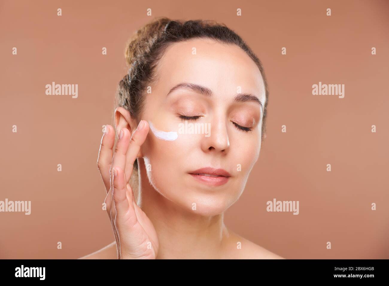 Jolie femme aux yeux fermés appliquant de la crème sur la zone sous-oculaire de son visage tout en prenant soin de la peau après l'hygiène du matin en isolement Banque D'Images