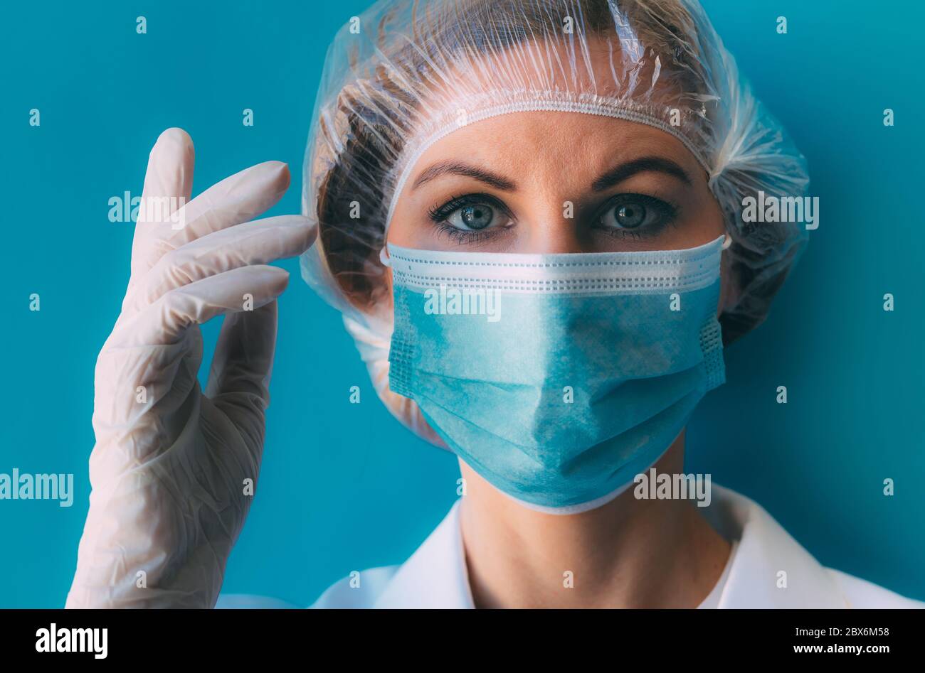 Portrait de jeune femme médecin en gros plan dans un capuchon médical, un  masque, une robe blanche et des gants sur fond bleu. Médecin avec la main  près de la tête sur