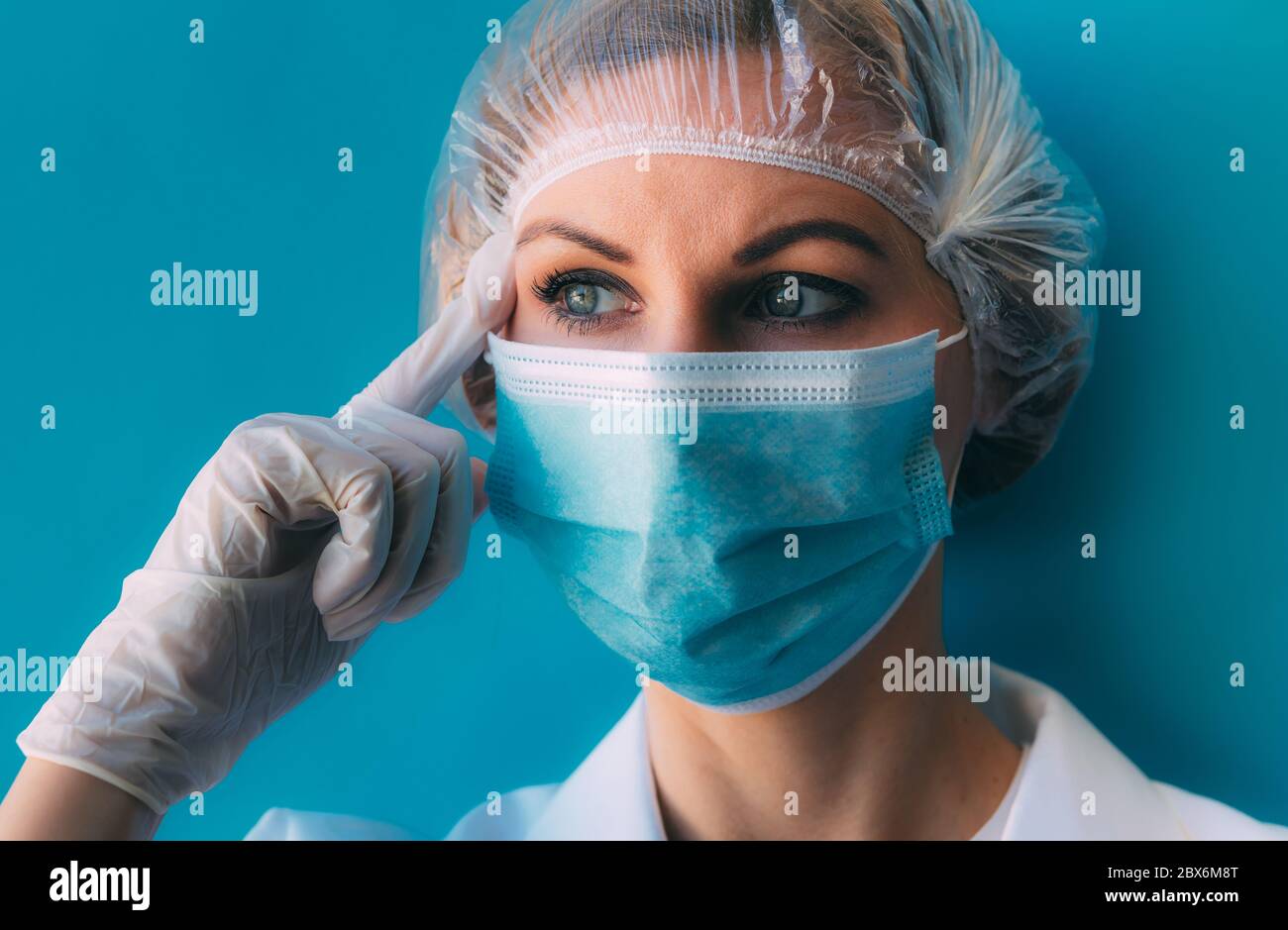 Portrait de jeune femme médecin en gros plan dans un capuchon médical, un masque, une robe blanche et des gants sur fond bleu. Médecin avec un doigt au temple, en pensant Banque D'Images