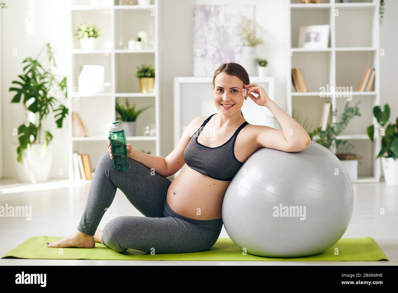 Jeune femme enceinte sportive avec succès, avec une bouteille d'eau reposant contre le fitball tout en étant assise sur un tapis dans un environnement domestique Banque D'Images