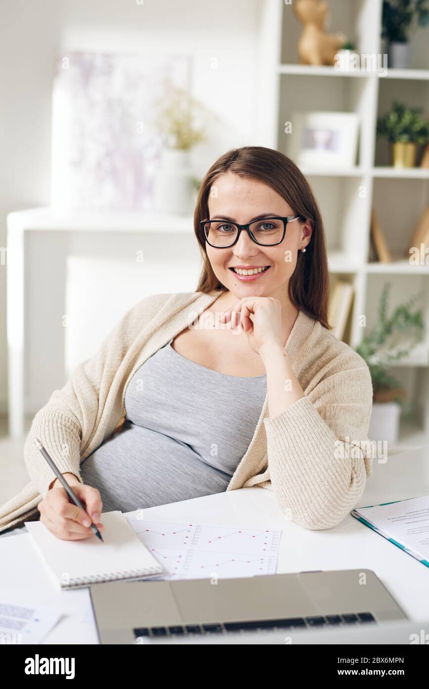 Jeune femme enceinte réussie analysant le graphique financier et prenant des notes dans le bloc-notes tout en travaillant devant l'appareil photo dans le bureau à domicile Banque D'Images