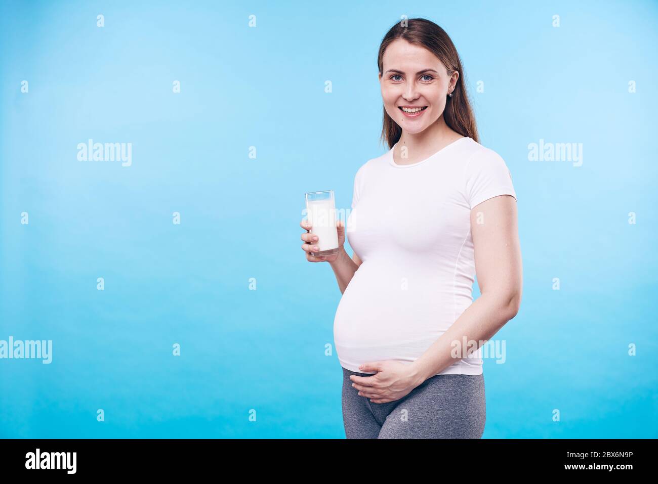 Bonne jeune femme avec un verre de lait touchant son ventre enceinte tout en disant que le calcium est nécessaire pour la future mère et son bébé Banque D'Images