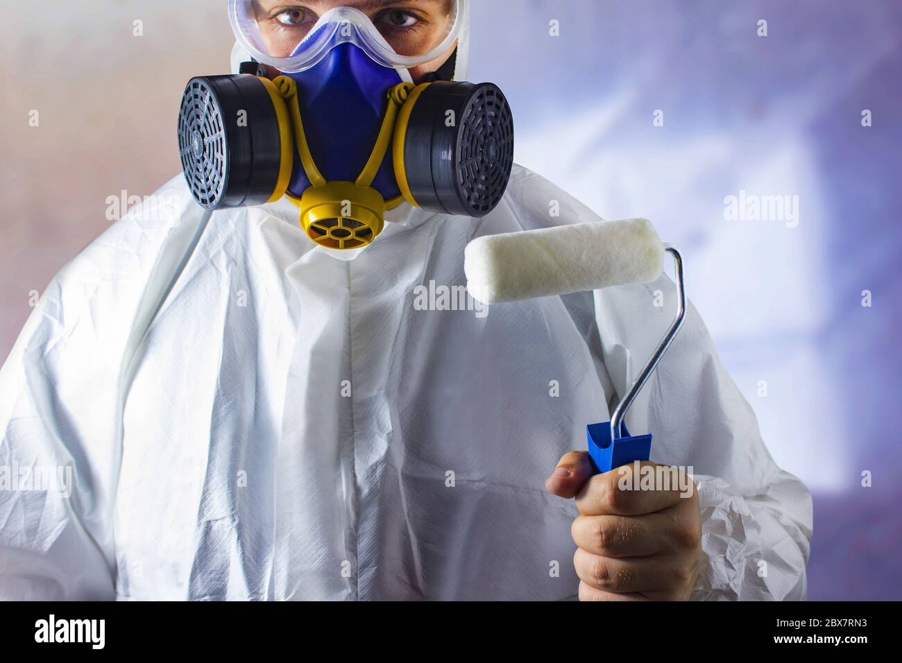 Travailleur en costume de protection et respirateur tenant un rouleau de peinture dans sa main vue rapprochée sur un arrière-plan abstrait flou. Banque D'Images