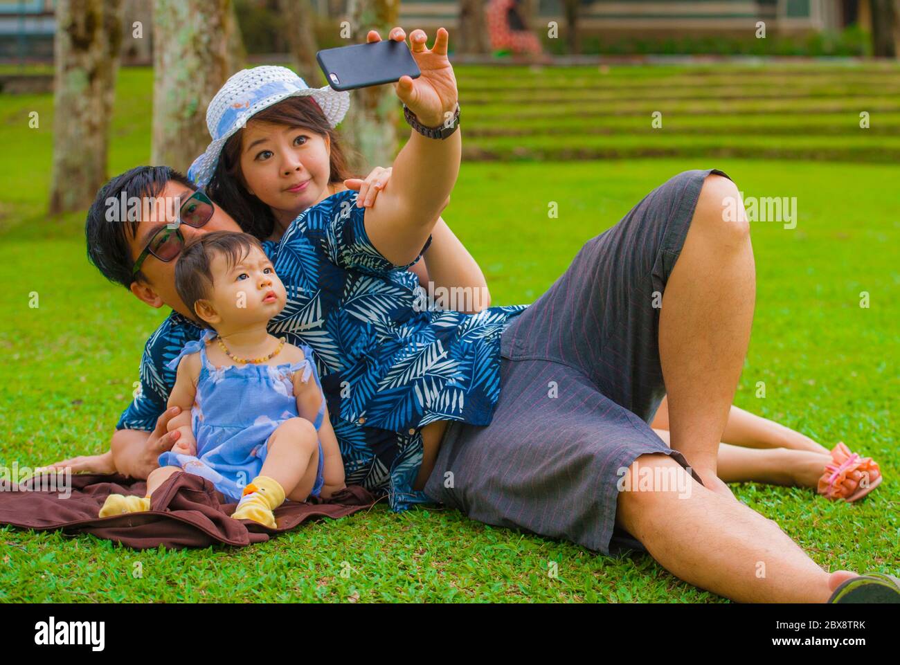Jeune famille coréenne asiatique heureuse et aimante avec parents et petite fille douce au parc de la ville avec le père prenant le selfie pic avec téléphone mobile Banque D'Images