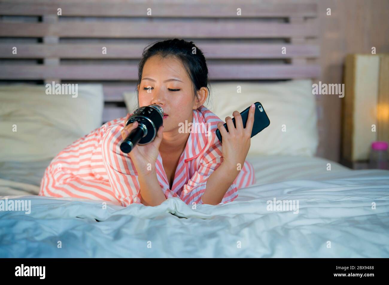 Jeune fille adolescente asiatique heureuse et belle, américaine chantant une chanson de karaoké excitée à la chambre à coucher de maison tenant le téléphone mobile jouant sur le lit excité et ch Banque D'Images