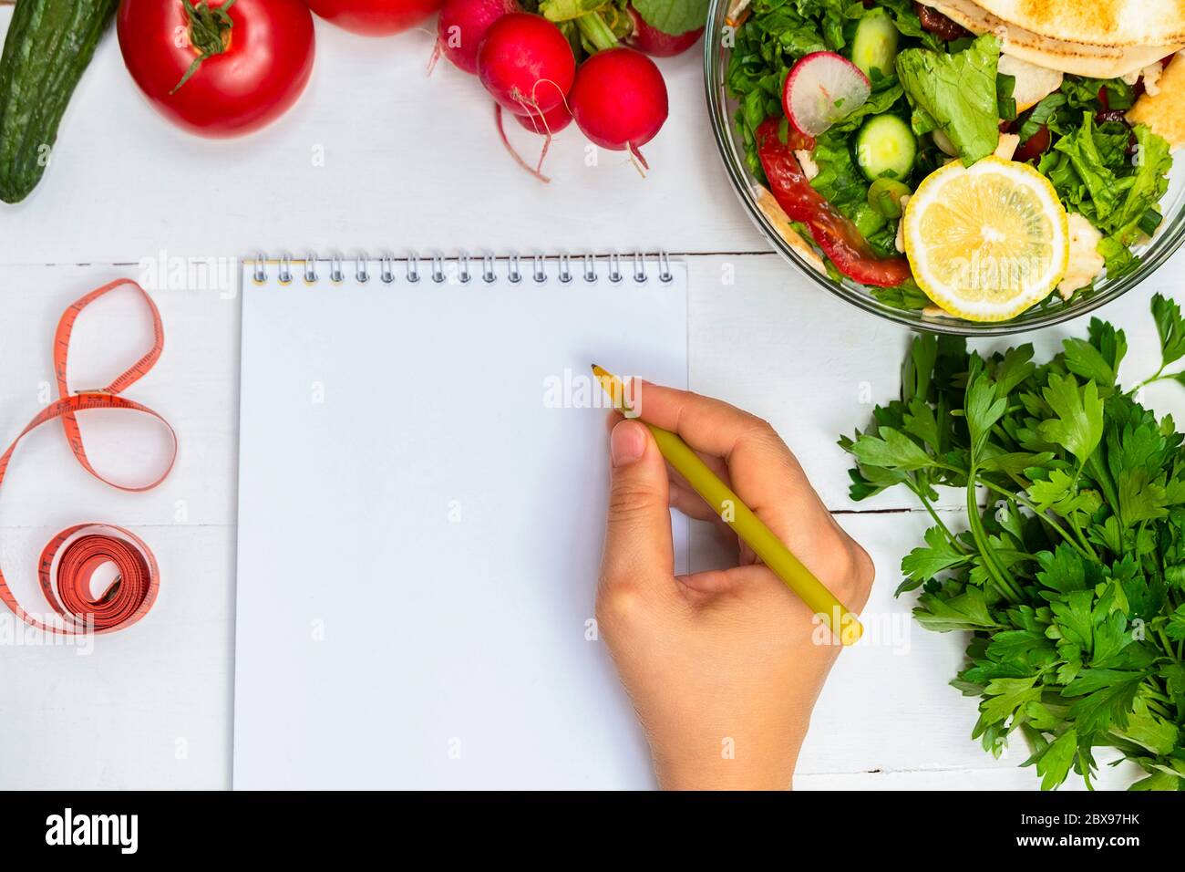 Vue de dessus du bloc-notes blanc vierge et du stylo main femme sur une table en bois blanc avec salade saine et légumes frais. Concept de régime alimentaire sain Banque D'Images