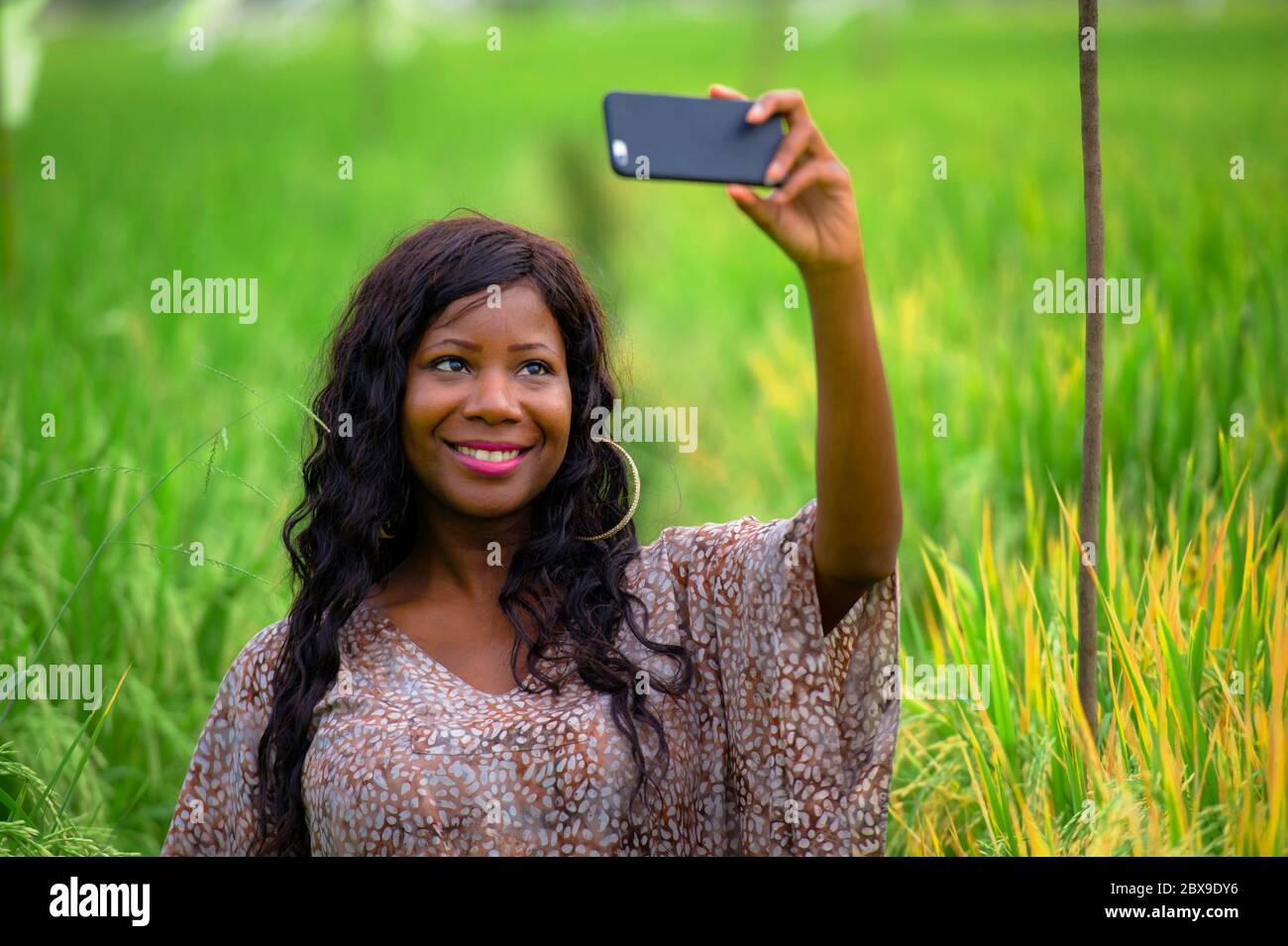 Jeune heureuse et belle noire afro-américaine dans la robe élégante posant cool prendre photo selfie avec téléphone mobile souriant gai rizieux champ Banque D'Images