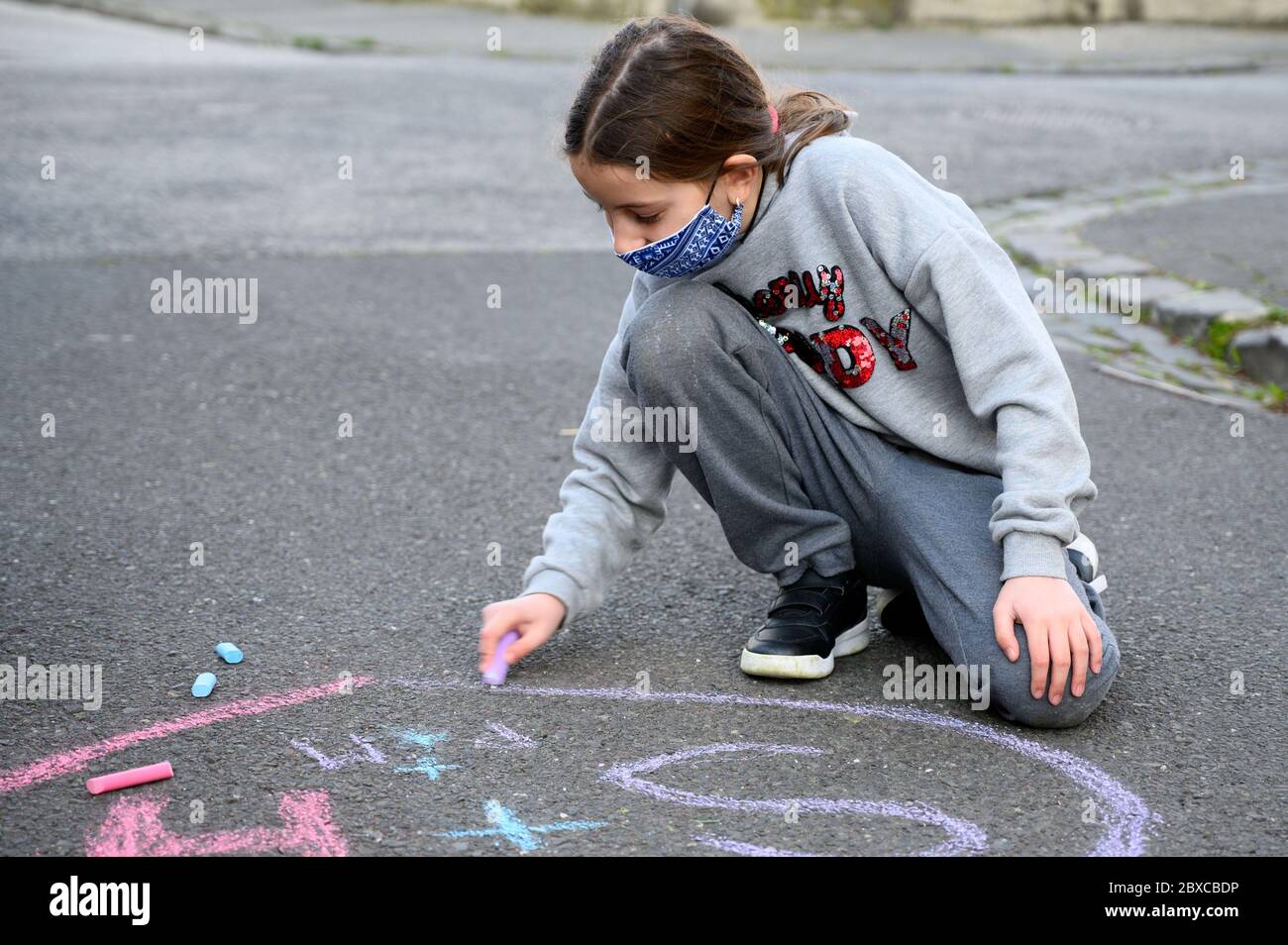 Une fille avec un masque de visage dessine un coeur sur le trottoir avec une craie. Elle porte un masque facial en raison de la pandémie du coronavirus. Banque D'Images