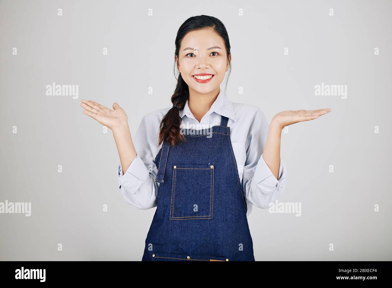 Portrait de jeune femme vietnamienne gaie en tablier en denim haussant les épaules Banque D'Images