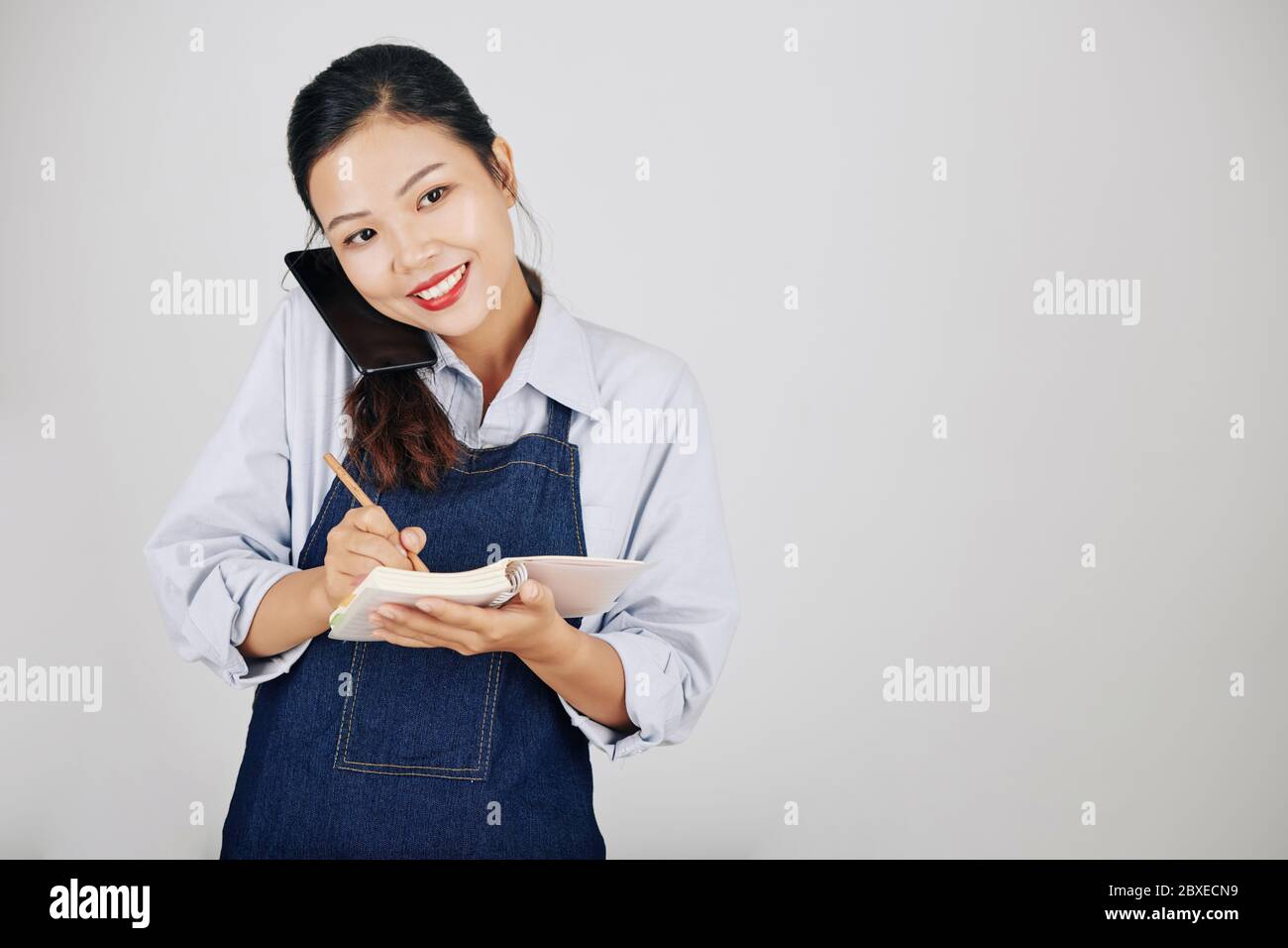 Souriant, jolie serveuse vietnamienne en tablier en denim parlant au téléphone avec le client et écrivant la commande Banque D'Images