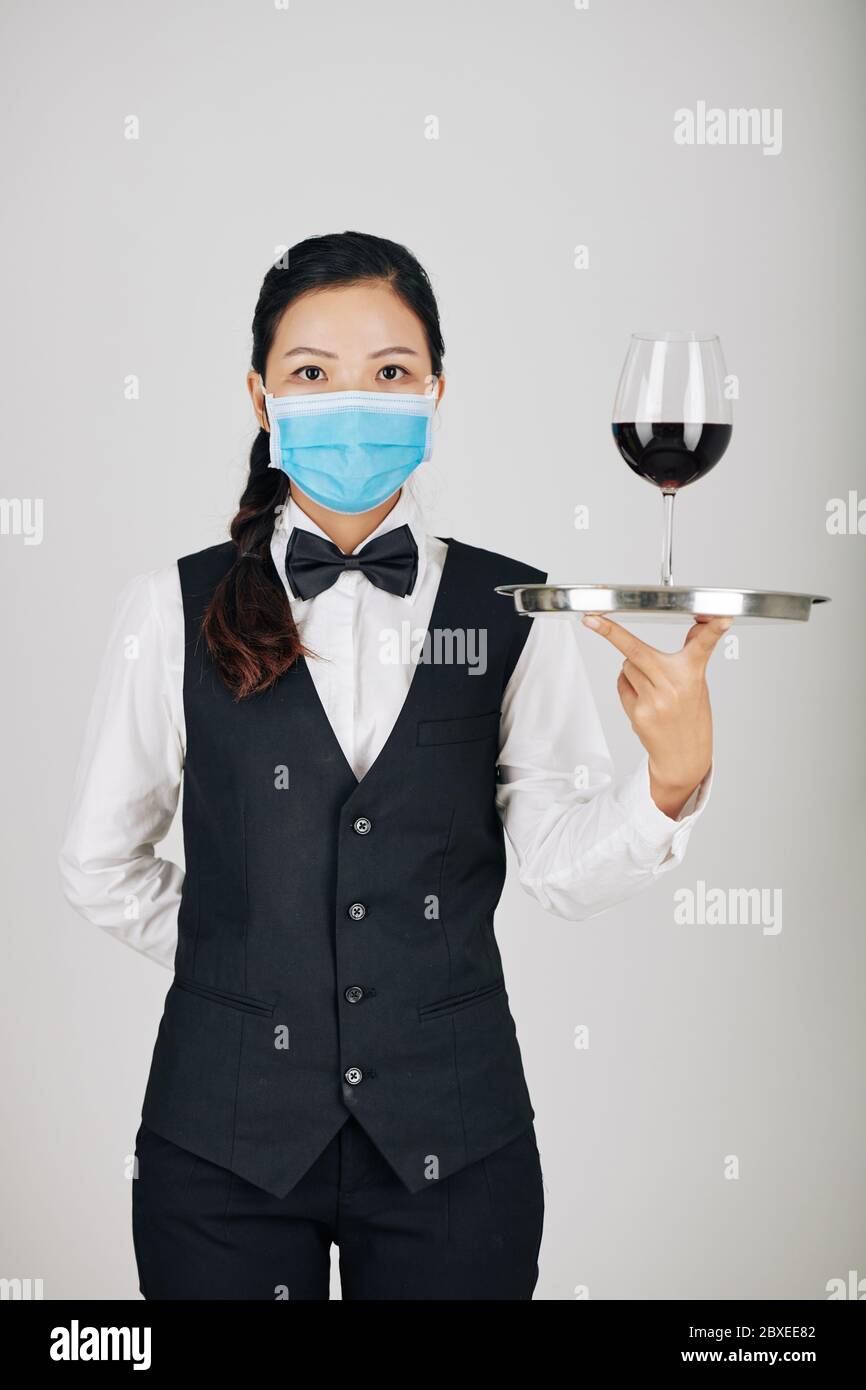 Jolie jeune serveuse vietnamienne dans un masque médical servant un verre de vin rouge Banque D'Images