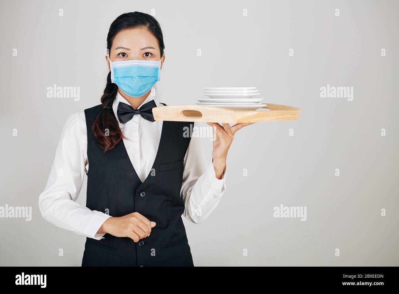 Portrait de jeune serveuse sérieuse dans un masque médical portant un plateau en bois avec pile de plaques Banque D'Images