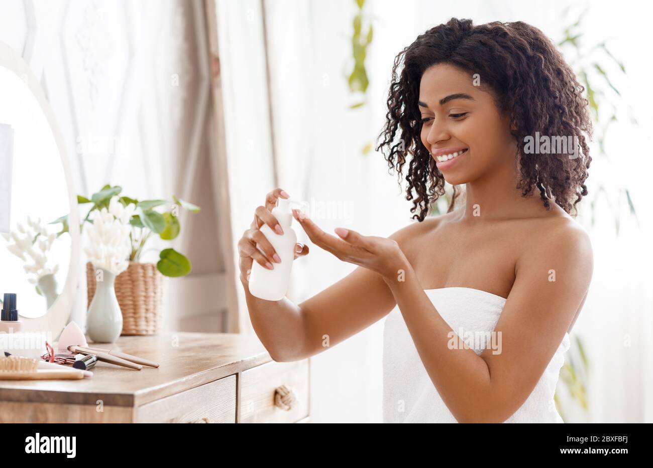 Hydratant la peau. Femme africaine souriante appliquant la lotion corporelle dans la chambre à la maison Banque D'Images