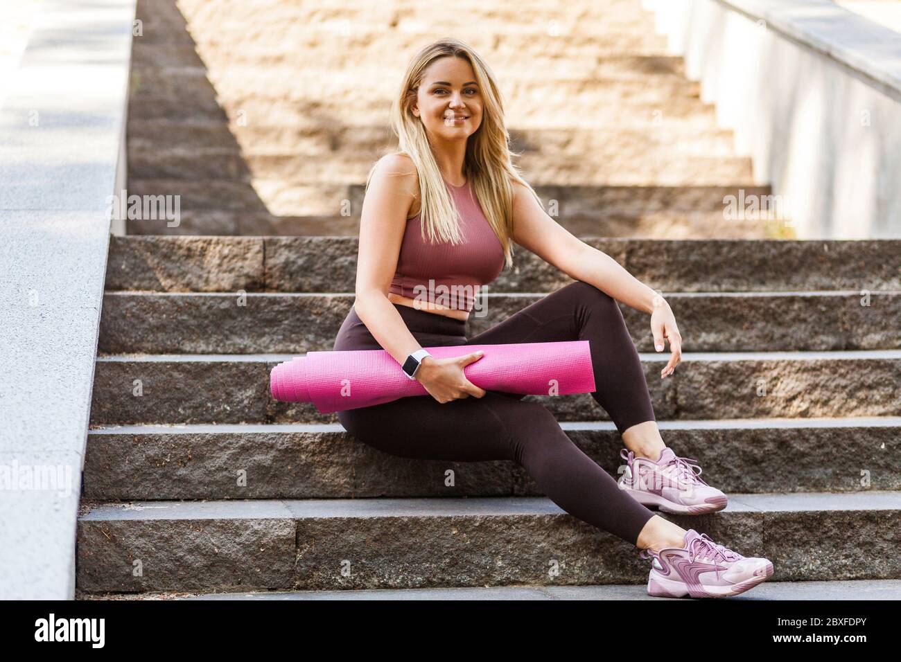 Bonne tenue blonde fille dans le pantalon de yoga assis sur les escaliers, tenant le tapis et souriant avec heureux expression satisfait, se détendre prendre une pause après le sport tra Banque D'Images