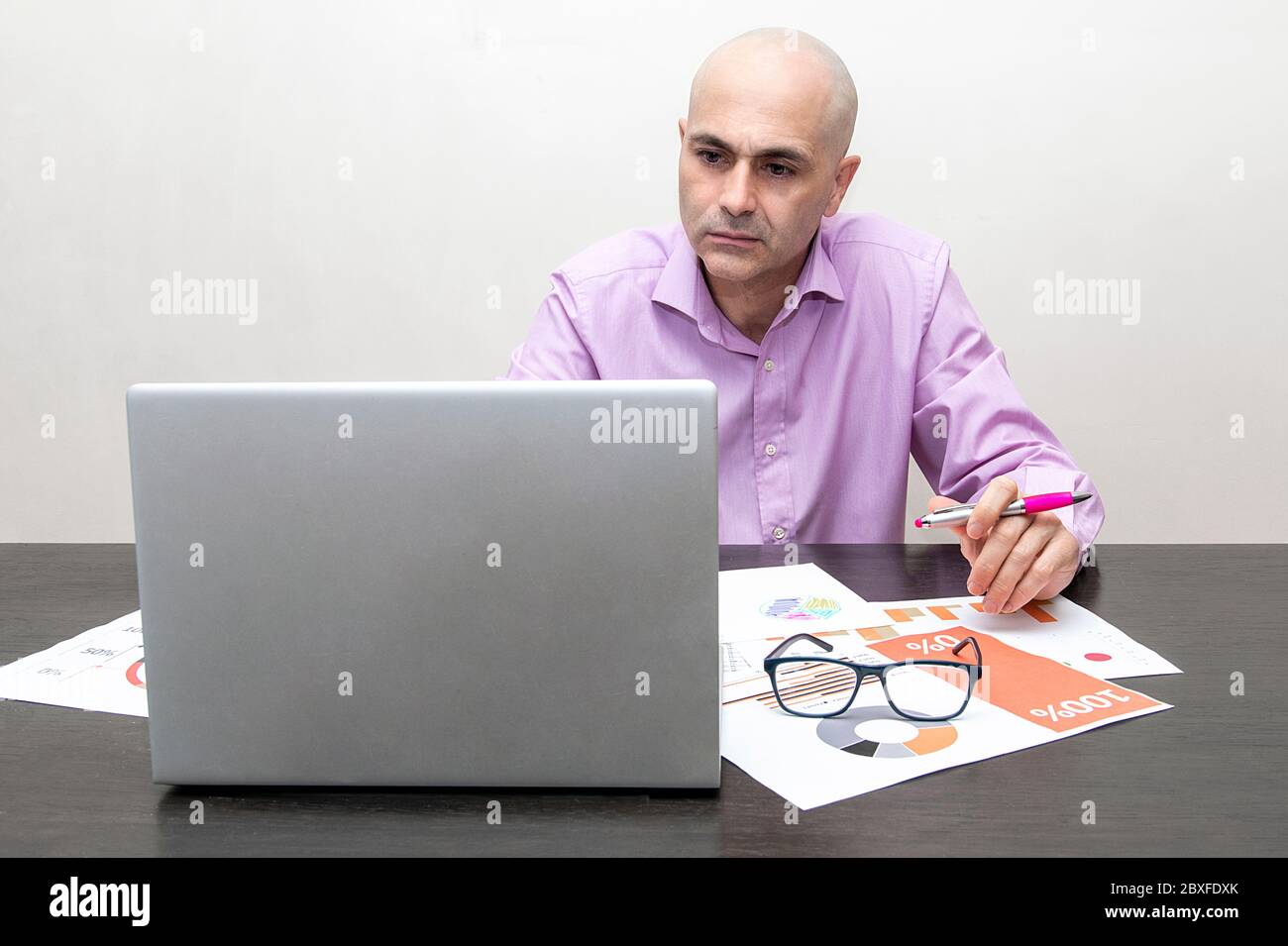 homme inquiet regardant un ordinateur portable avec un stylo dans sa main, des lunettes et des papiers avec des graphiques Banque D'Images