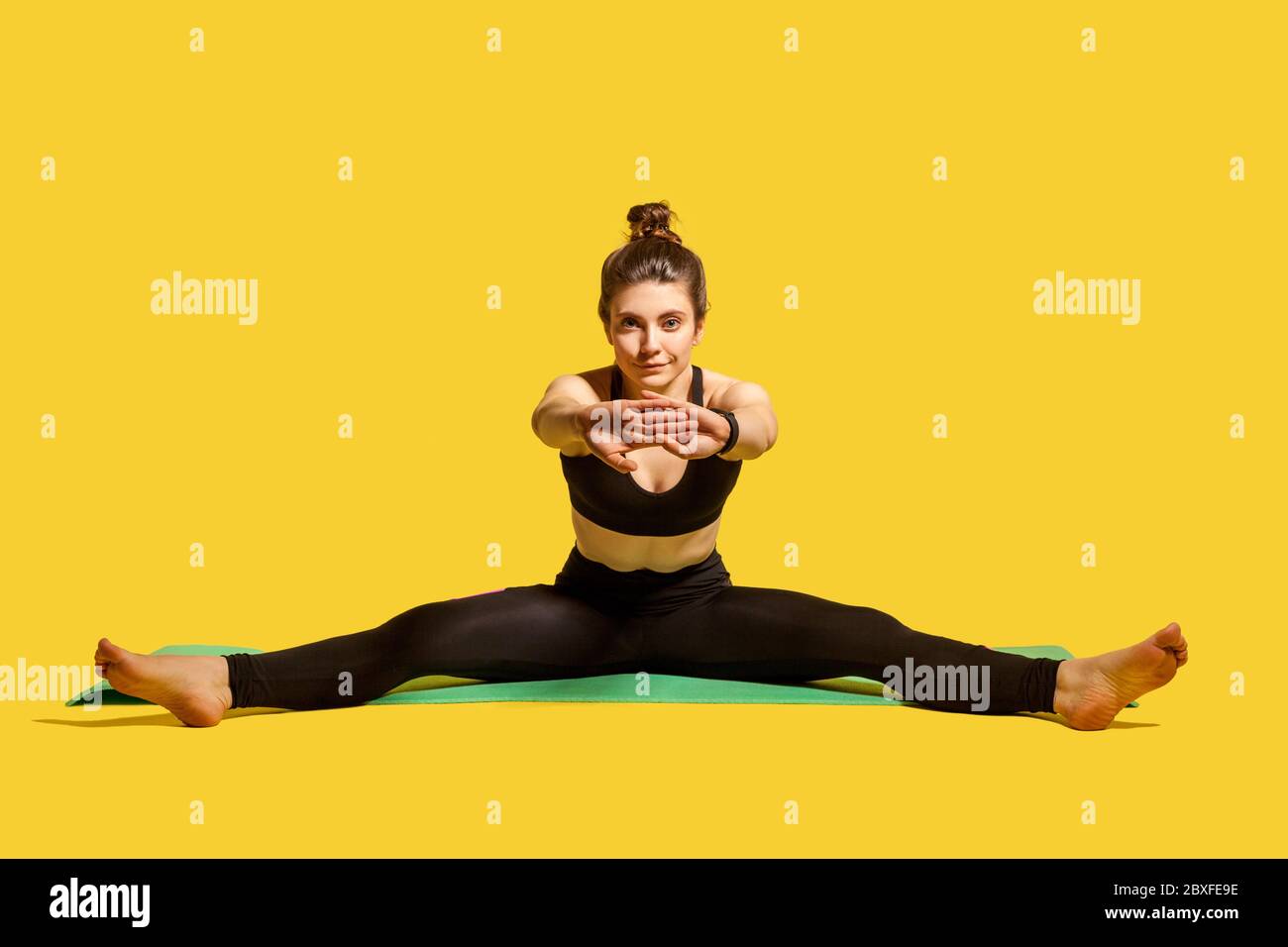 Mettre la femme de gymnastique avec un pain de cheveux dans les vêtements de sport serrés assis sur le tapis avec les jambes et les mains étirant, en se réchauffant, faisant des exercices de flexibilité sportive Banque D'Images