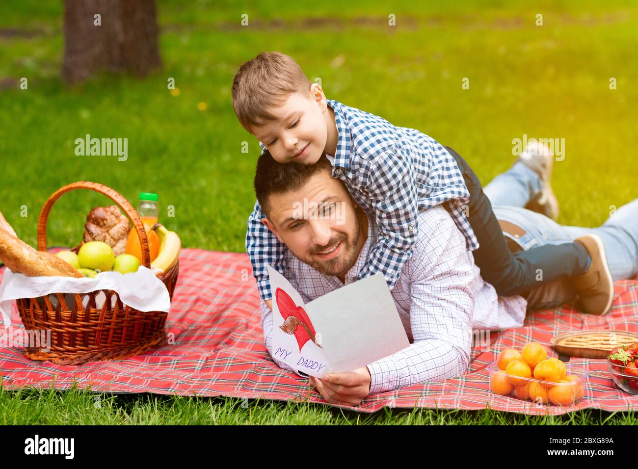Bonne journée du Père. Un enfant mignon embrassant son père, pendant qu'il lit une carte de vœux sur un pique-nique en forêt Banque D'Images