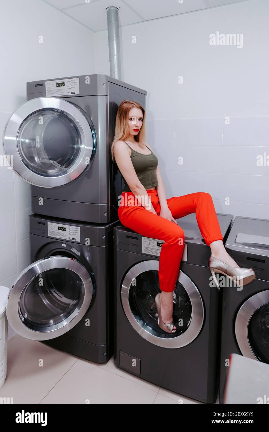 Jolie femme en blanchisserie se trouve sur et contre les machines à laver ; elle porte un pantalon rouge vif, des talons hauts et un haut ; ses lèvres sont rouges vif Banque D'Images