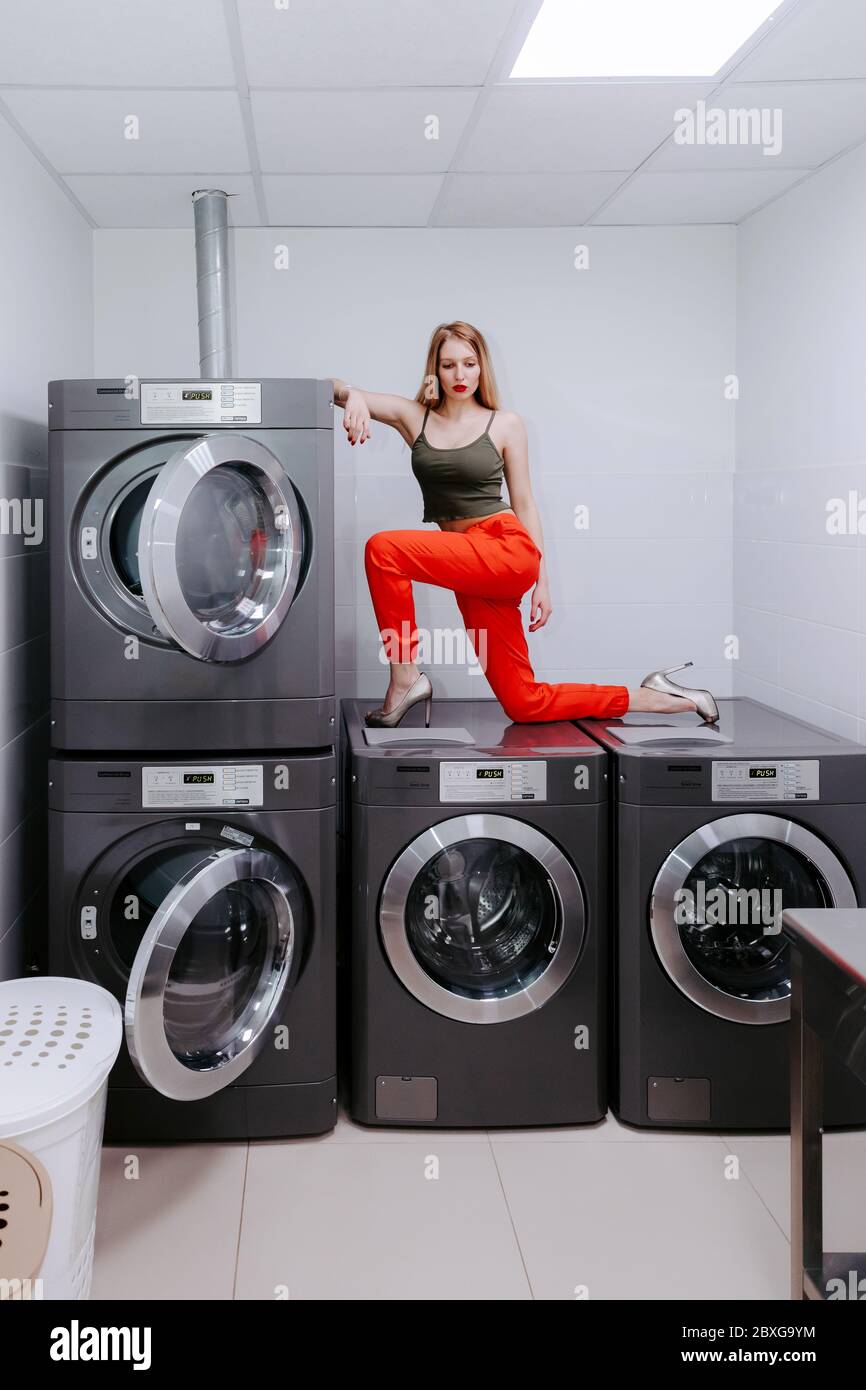 Jolie femme en blanchisserie est debout sur plusieurs machines à laver, elle porte un pantalon rouge vif, des talons hauts et un haut, ses lèvres sont rouge vif. Banque D'Images