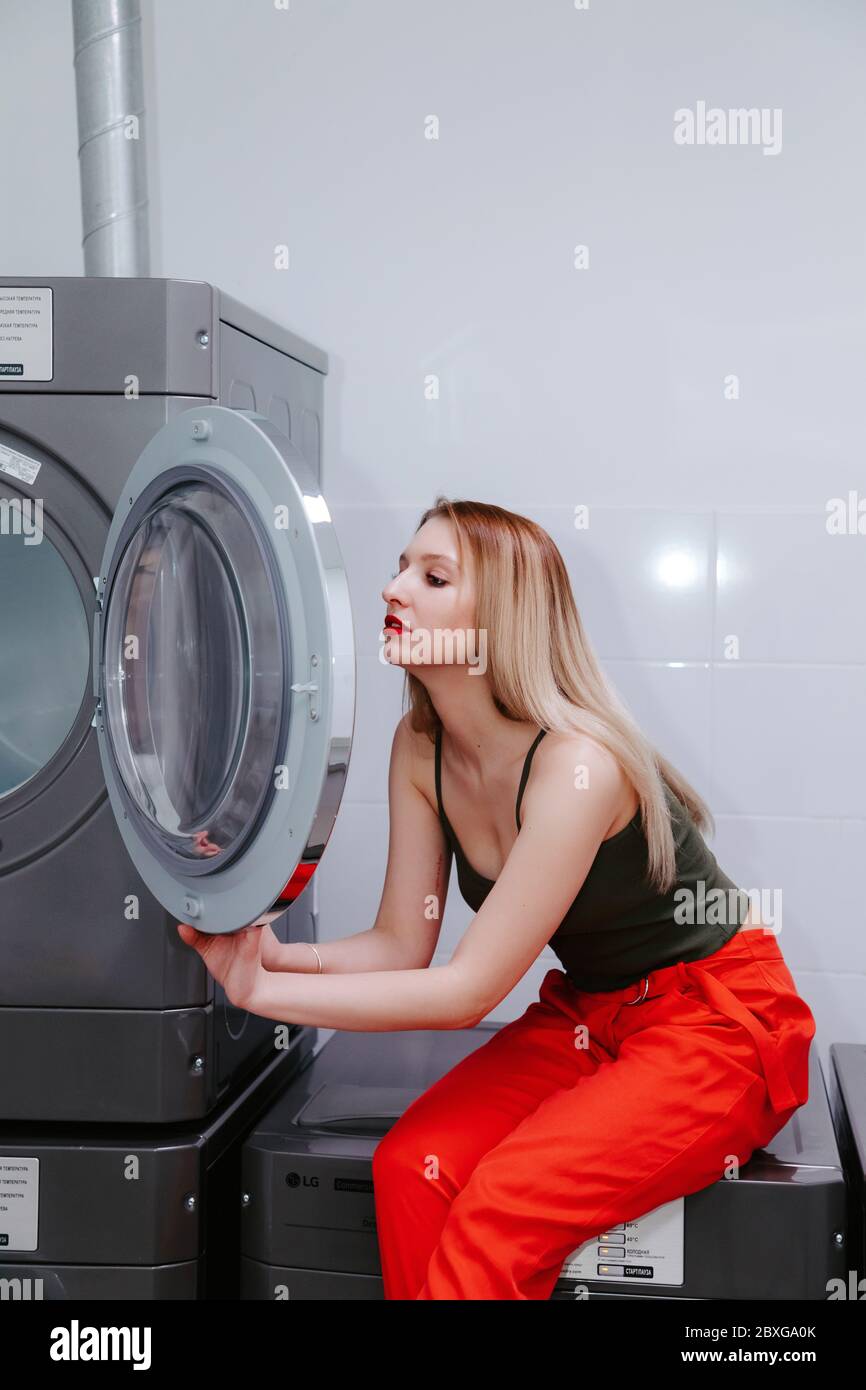 Jolie femme se regarde dans la porte vitrée d'une machine à laver, elle porte un pantalon rouge vif et un haut, ses lèvres sont rouge vif. Banque D'Images