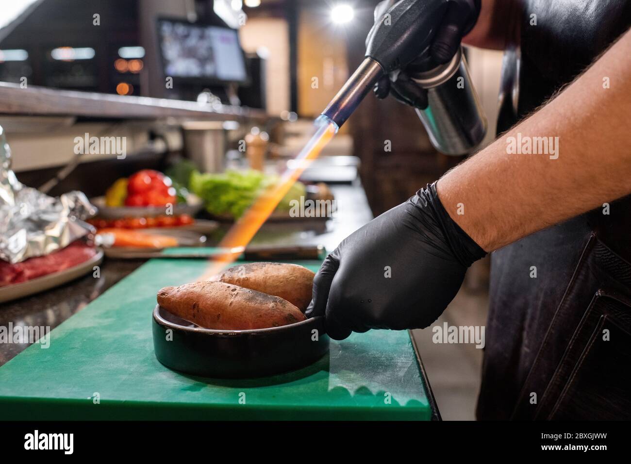 Gros plan d'un chef méconnaissable avec des gants noirs et une lampe à gaz pour cuire des patates douces dans la cuisine du restaurant Banque D'Images