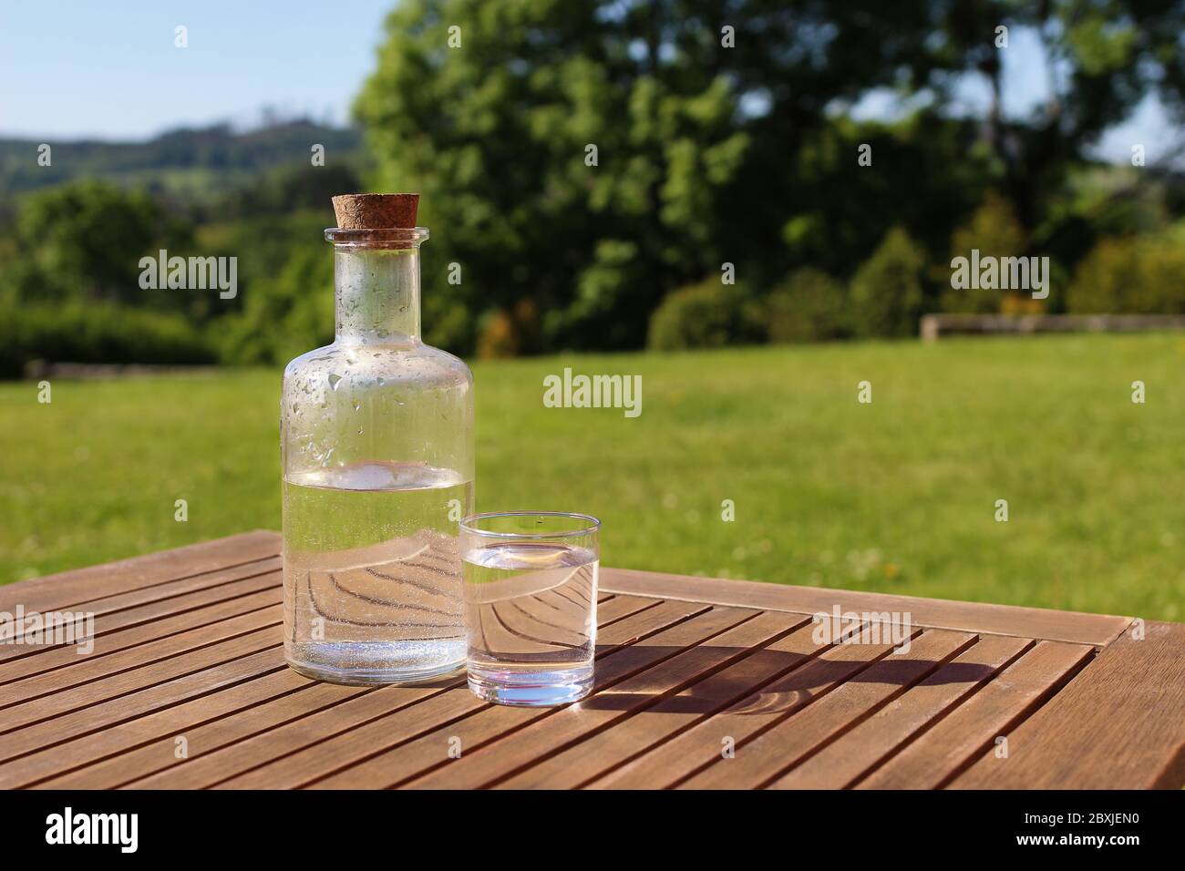 Bouteille et verre d'eau sur table en bois avec chaise. Arbres verts flous  et sommet de colline arrière-plan. Petit déjeuner en plein air dans la  campagne. Pique-nique, jardin p Photo Stock -