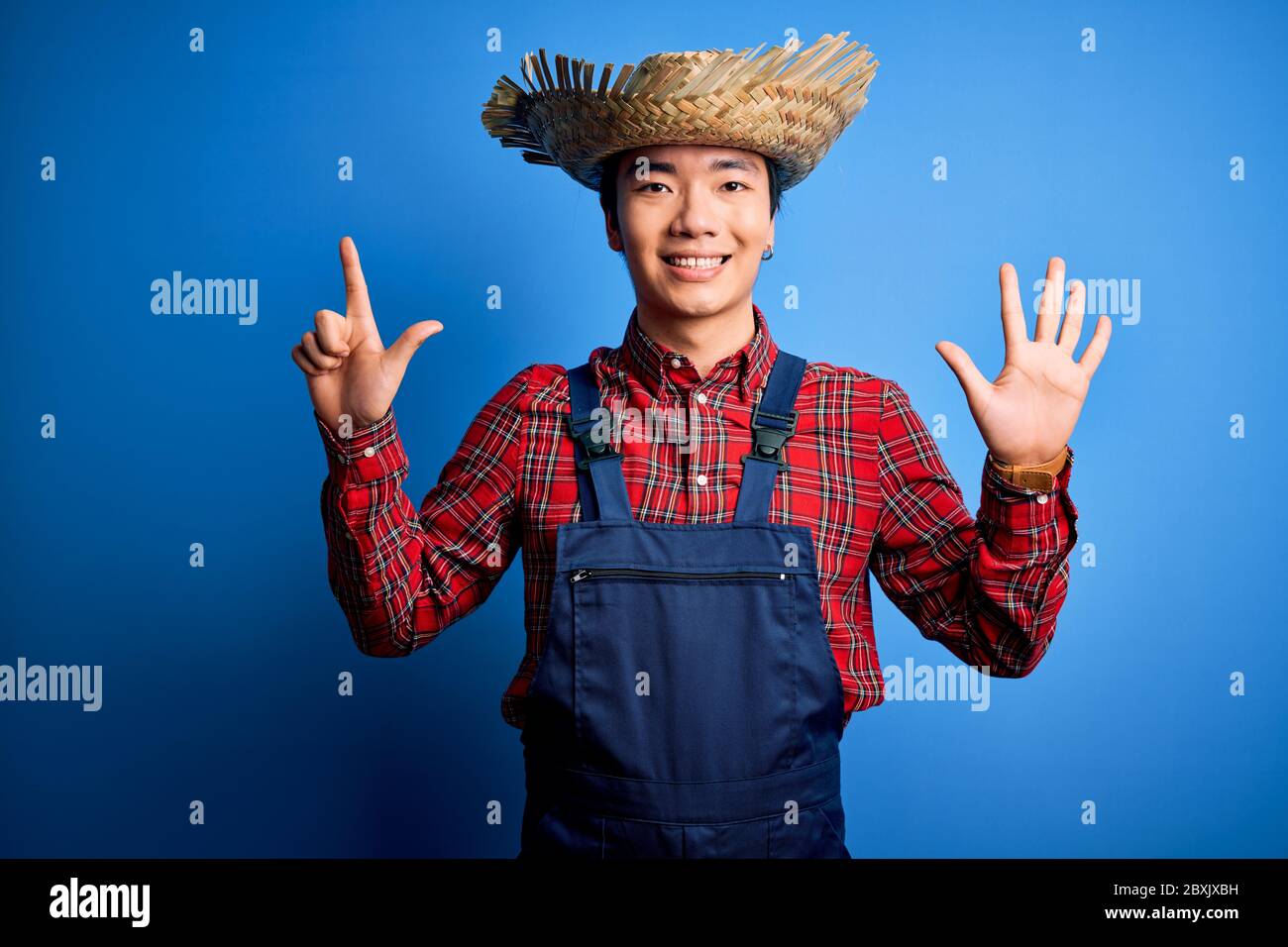 Jeune fermier chinois charmant portant un tablier et chapeau de paille sur fond bleu montrant et pointant vers le haut avec les doigts numéro sept tout en souriant con Banque D'Images